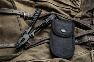 A multi-tool with pliers is placed on a brown canvas bag with leather straps, accompanied by a black nylon sheath, showcasing its utility and rugged design photo