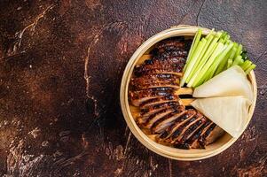 Peking Duck in bamboo steamer served with cucumber, green onion, and wheaten chinese pancakes. Dark background. Top view. Copy space photo