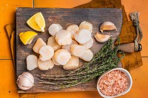 Uncooked Raw sea scallops fillets on a wooden board with thyme and lemon. Orange background. Top view photo