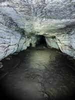 Deep within a dark and enigmatic cave, an intriguing light shines down from the ceiling, casting illumination upon the unusual rock formations and the surrounding landscape all around photo