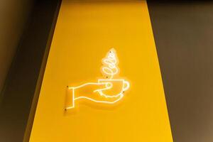 A neon sign on a yellow wall displays a hand with a coffee cup photo
