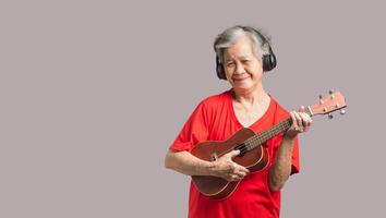 Portrait of an elderly Asian woman with short gray hair playing the ukulele with a smile while standing on a gray background photo