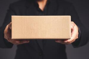 Businessman in a black suit holds and points to a cardboard box while standing on a gray background photo