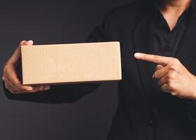 Businessman in a black suit holds and points to a cardboard box while standing on a gray background photo