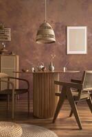 Stylish boho vintage dining room interior with poster mock up. Round table with rattan armchair, round carpet with pouf, vase with dried flowers and cup. Brown wall and rattan lamp. Mock up poster. photo