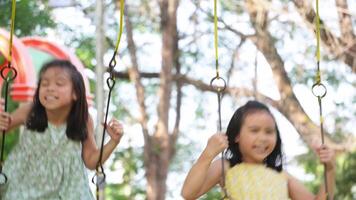 Cute sisters are having fun playing on the swings in a playground in a park. Children playing in the playground during summer vacation. video