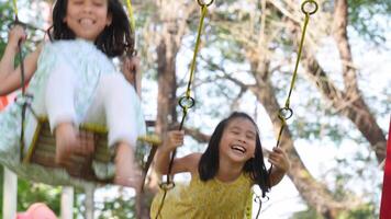 Cute sisters are having fun playing on the swings in a playground in a park. Children playing in the playground during summer vacation. video