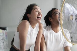 Two young Asian startup coworkers are fun blowing wind with electric fan, working in hot temperatures in a casual small business office in summer, refreshing from uncomfortable heat weather season. photo