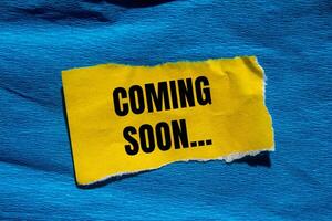 Coming soon message written on ripped torn yellow paper piece with blue background. Conceptual coming soon symbol. Copy space. photo