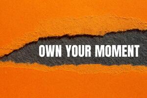 Own your moment message written on ripped torn orange paper with gray background. Conceptual own your moment symbol. Copy space. photo