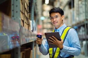 A man wearing a yellow vest and glasses is standing in a warehouse with a tablet in his hand. He is using a barcode scanner to scan items photo