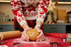 Woman in cozy red sweater prepares gingerbread dough for baking holiday cookies in kitchen. Close-up of hands kneading dough on red mat surrounded by cookie cutters and Christmas decorations photo