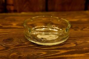 Clear glass ashtray sits on a rustic wooden table with remnants of tobacco at a casual gathering spot in the evening photo