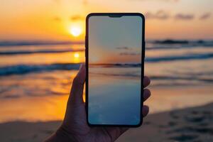 A person holds a smartphone featuring a blank display as a stunning sunset illuminates the beach. The golden hues reflect on the water, creating a serene atmosphere photo