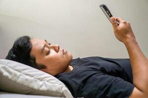 Young Asian man lying on bed while using smart phone. Man using smartphone in bed at night. Bad habit concept. photo