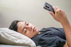 Young Asian man lying on bed while using smart phone. Man using smartphone in bed at night. Bad habit concept. photo