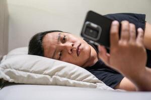 Young Asian man lying on bed while using smart phone. Man using smartphone in bed at night. Bad habit concept. photo