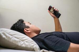 Young Asian man lying on bed while using smart phone. Man using smartphone in bed at night. Bad habit concept. photo