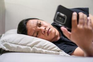 Young Asian man lying on bed while using smart phone. Man using smartphone in bed at night. Bad habit concept. photo