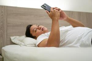 Young Asian man lying on bed while using smart phone. Man using smartphone in bed at night. Bad habit concept. photo