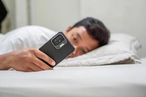Young Asian man lying on bed while using smart phone. Man using smartphone in bed at night. Bad habit concept. photo