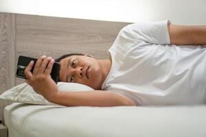 Young Asian man lying on bed while using smart phone. Man using smartphone in bed at night. Bad habit concept. photo