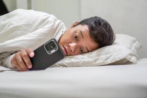 Young Asian man lying on bed while using smart phone. Man using smartphone in bed at night. Bad habit concept. photo
