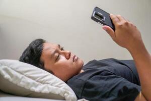 Young Asian man lying on bed while using smart phone. Man using smartphone in bed at night. Bad habit concept. photo