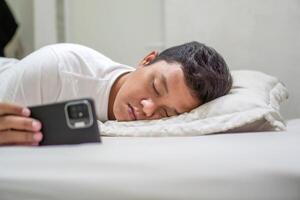 Tired Asian man fall asleep on bed while using mobile phone. Man sleep in bed at night and still holding his phone. Bad habit concept. photo