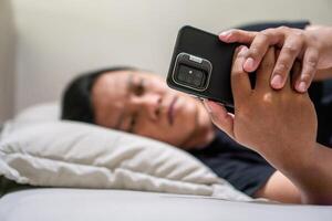 Young Asian man lying on bed while using smart phone. Man using smartphone in bed at night. Bad habit concept. photo