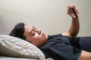 Young Asian man lying on bed while using smart phone. Man using smartphone in bed at night. Bad habit concept. photo