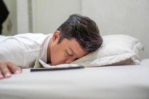 Tired Asian man fall asleep on bed while using mobile phone. Man sleep in bed at night and still holding his phone. Bad habit concept. photo
