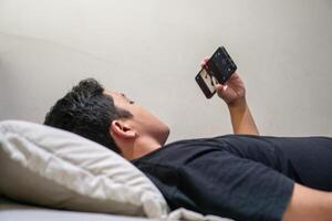 Young Asian man lying on bed while using smart phone. Man using smartphone in bed at night. Bad habit concept. photo