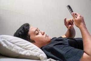 Young Asian man lying on bed while using smart phone. Man using smartphone in bed at night. Bad habit concept. photo