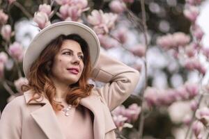 Woman magnolia flowers, surrounded by blossoming trees., hair down, white hat, wearing a light coat. Captured during spring, showcasing natural beauty and seasonal change. photo