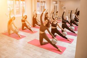 A group of six athletic women doing pilates or yoga on pink mats in front of a window in a beige loft studio interior. Teamwork, good mood and healthy lifestyle concept. photo