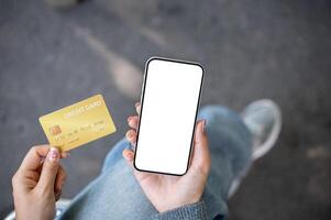 A top view of a woman sitting outdoors, holding a credit card and a smartphone with a screen mockup. photo