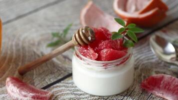 homemade sweet yogurt in a glass jar with pieces of red ripe grapefruit and honey, on a wooden table video