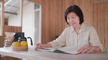 Asian senior woman sitting reading a book calmly and relaxed on a chair on a table at home in bamboo, coffee dripper and kettle and lemon on the table, searching for information, novel and knowledge video