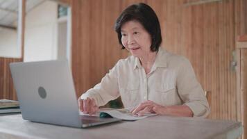 Close-up of senior woman talking on call using laptop and reading book on table in bamboo house. Online learning. call with family. Searching for information. Seniors learning online. video
