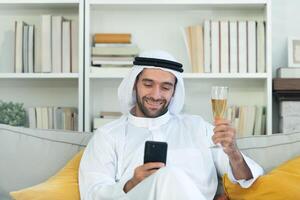 Portrait of a smiling Arabian man holding a glass of champagne and using mobile phone at home photo