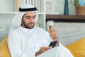 Portrait of a smiling Arabian man holding a glass of champagne and using mobile phone at home photo
