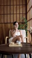 Young, caucasian woman talking on the phone, drink cup of tea lounge chilling near window in modern cozy apartment. Relaxed brunette girl feel fatigue lounge on comfortable sofa at home. Vertical shot video