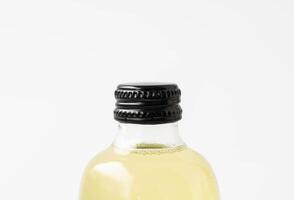 Small transparent glass bottle with black cap and white label, filled with colorful kombucha of yellow, standing on a white surface. photo