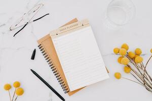 Flat lay of white notebook with copy space for text, glasses and yellow flowers on white marble background photo