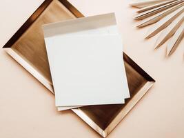 White blank paper square card mockup, envelope on gold tray and dry palm leaf on beige background photo