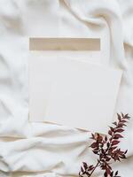 White blank paper sheet card mockup, envelope and dry floral branch on neutral white textile background. photo