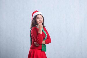 portrait of beautiful girl wearing christmas sweater, santa hat and scarf, slanted left smile to camera hand sign pointing to right, isolated on white background photo