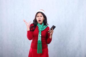 young asian woman surprised at camera holding mobile phone while growling,wearing red sweater with Santa hat and scarf celebrating merry christmas and new year,isolated on white background photo