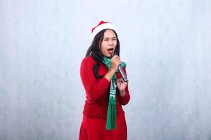 portrait of Asian female wearing red sweater with Santa hat and scarf celebrating merry christmas, shouting singing closing eyes both hands holding karaoke mic, isolated on white background photo